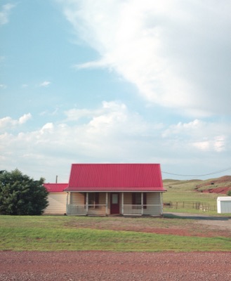 Lone red-roofed farmhouse standing on a quiet stretch of red-dirt prairie under a wide sky