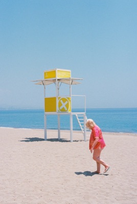 Woman in a red blouse walking past a yellow lifeguard tower on an empty Mediterranean beach