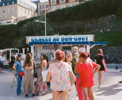 A crowd gathered around the 'Glaces au Bon Goût' kiosk on a sunny Basque promenade