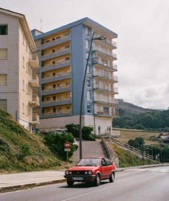 Red Volkswagen Polo passing a curved blue-tiled apartment block in a Basque hill town