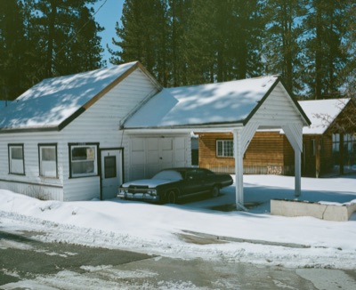 Snow-covered cabin with a vintage sedan parked under the carport in pine country