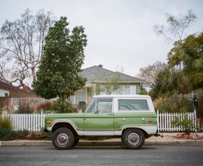 Restored green Ford Bronco parked outside a white picket-fenced bungalow